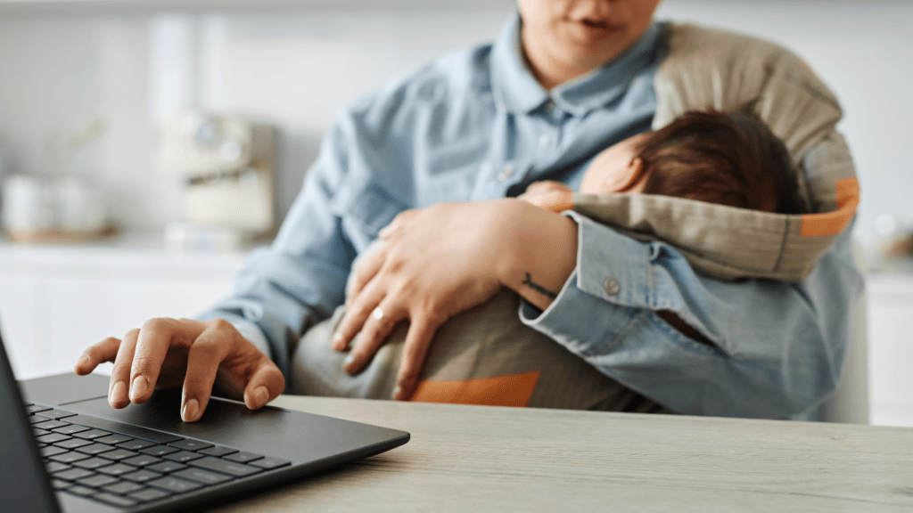 Someone holding a baby while sitting at a desk with a laptop